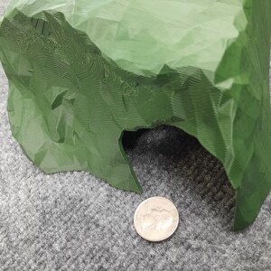 Pode incluir: Uma caverna verde impressa em 3D com uma superf&iacute;cie texturizada e uma pequena abertura. A caverna est&aacute; sobre uma superf&iacute;cie cinza.