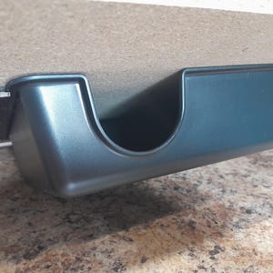 May include: A matte black under-desk storage tray with a smooth, rectangular design and a large U-shaped cutout on the front edge. The tray is securely mounted to the underside of a flat surface using a black bracket and visible metal screws.
