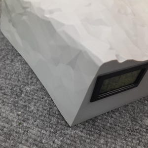 May include: A white, rock-shaped digital clock with a black display. The clock is on a gray surface.
