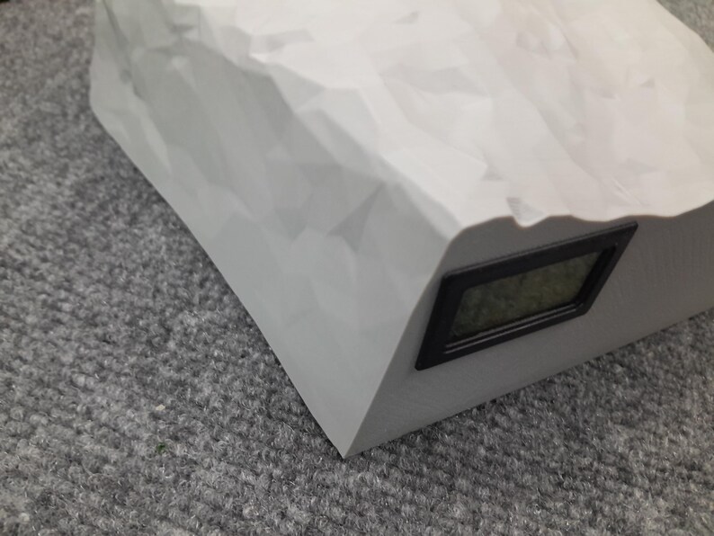 May include: A white, rock-shaped digital clock with a black display. The clock is on a gray surface.