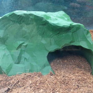 Pode incluir: Um esconderijo de pl&aacute;stico verde para r&eacute;pteis, com a forma de uma rocha, com uma superf&iacute;cie texturizada. O esconderijo &eacute; projetado para fornecer um espa&ccedil;o seguro e protegido para os r&eacute;pteis se esconderem e descansarem.