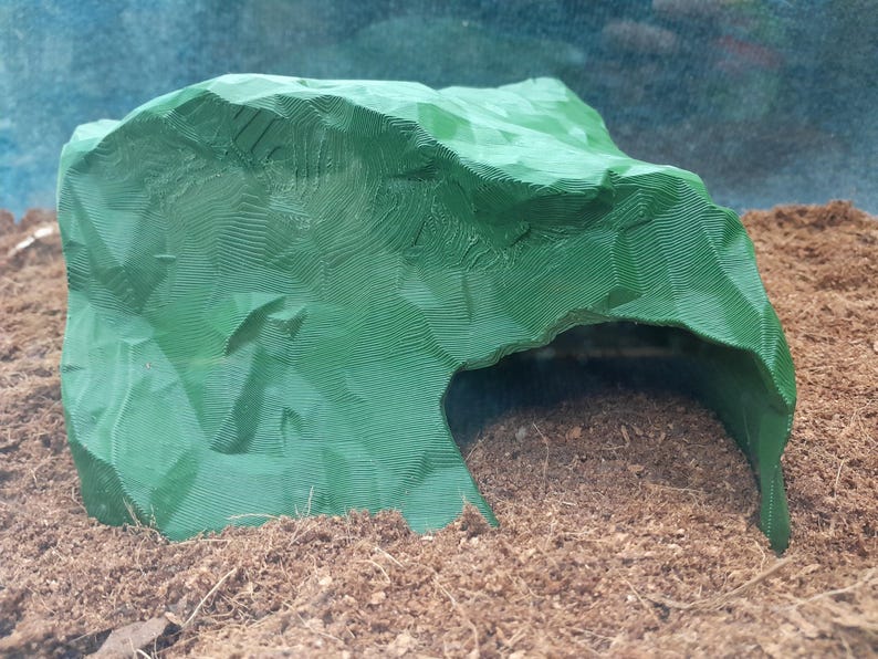 Pode incluir: Um esconderijo de pl&aacute;stico verde para r&eacute;pteis, com a forma de uma rocha, com uma superf&iacute;cie texturizada. O esconderijo &eacute; projetado para fornecer um espa&ccedil;o seguro e protegido para os r&eacute;pteis se esconderem e descansarem.