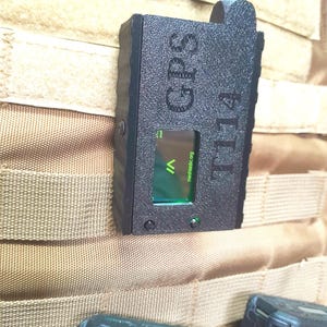 May include: Black GPS device labeled "GPS T14" with a small green screen displaying a graphic and the text "meshtastic.org". The device has a textured surface and a black antenna with a gold connector. It is attached to a tan fabric.