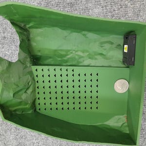 Pode incluir: Um recipiente de pl&aacute;stico verde com um inserto de padr&atilde;o triangular e um pequeno dispositivo eletr&ocirc;nico preto. O recipiente tem uma aba que pode ser levantada para acessar o interior.