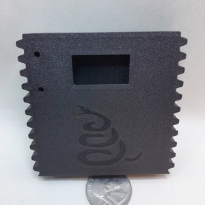 May include: Black plastic box with a cutout for a display screen and a snake logo. The box has a textured surface and a serrated edge.