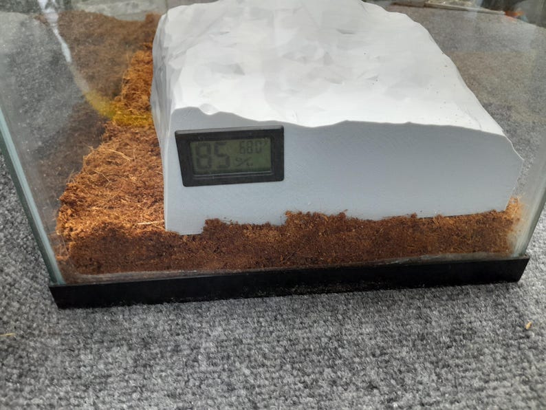 May include: A white, rock-like structure with a digital thermometer attached to the side. The thermometer reads 85.6 degrees. The structure is sitting on a brown, shredded coconut substrate in a glass terrarium.