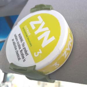 May include: A round white and yellow Zyn Citrus nicotine pouch container held by a matte olive green plastic clip. The label features the text ZYN, Citrus, 15 nicotine pouches, 3 mg, and a warning about nicotine being an addictive chemical.