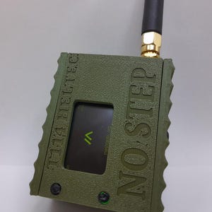 May include: A green, rectangular device with a black antenna and a small screen displaying the text "meshmastic.org". The device is labeled "MO.S.T.E.R.  GET TIME BACK".