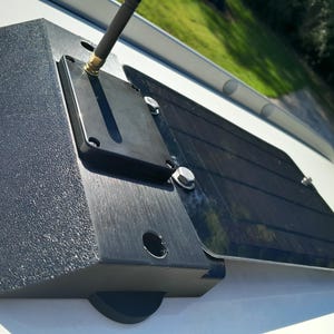 May include: A black solar panel system with a textured, dark gray base and a hinged, rectangular solar panel. A black antenna extends from a mounted component. The system is secured with silver bolts and is mounted on a white surface.