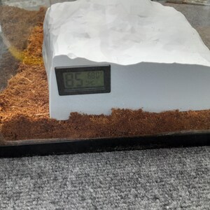 May include: A white, rock-like structure with a digital thermometer attached to the side. The thermometer reads 85.6 degrees. The structure is sitting on a brown, shredded coconut substrate inside a glass terrarium.
