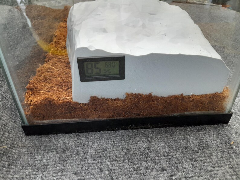 May include: A white, rock-like structure with a digital thermometer attached to the side. The thermometer reads 85.6 degrees. The structure is sitting on a brown, shredded coconut substrate inside a glass terrarium.