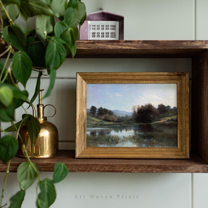 May include: A framed landscape painting of a lake with trees and a blue sky. The painting is in a wooden frame with a gold finish. The frame is sitting on a wooden shelf.