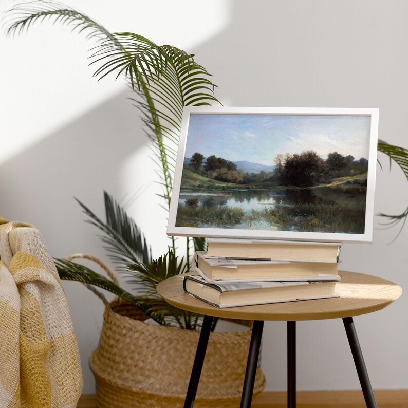 May include: A white framed landscape painting of a lake with trees and mountains in the background. The painting is sitting on a stack of books on a wooden table.