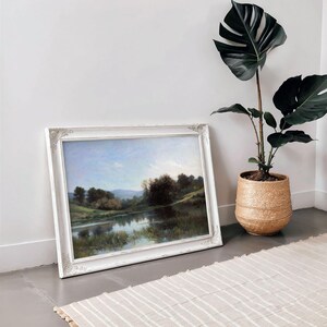 May include: A framed landscape painting of a lake with trees and hills in the background. The frame is white with a decorative design. The painting is hanging on a white wall next to a potted plant.