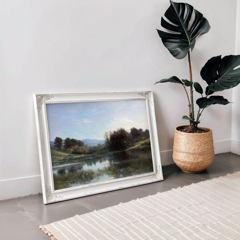 May include: A framed landscape painting of a lake with trees and hills in the background. The frame is white with a decorative design. The painting is hanging on a white wall next to a potted plant.