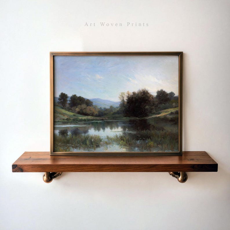 May include: A framed landscape painting of a lake surrounded by trees and hills. The painting is in a gold frame and is hanging on a wooden shelf with brass brackets.