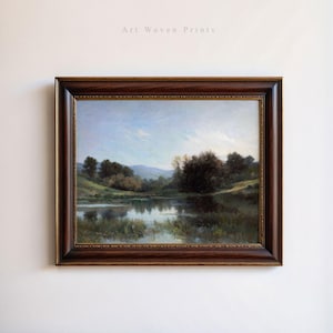 May include: A framed painting of a landscape with a lake in the foreground, surrounded by trees and hills. The sky is blue with white clouds. The painting is in a brown wooden frame.
