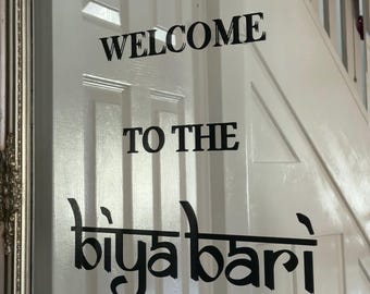 May include: Black vinyl lettering applied to a white panelled door. The text reads WELCOME TO THE in a standard sans-serif font, followed by the word biya bari in a stylized, decorative script font with a horizontal line running through it.
