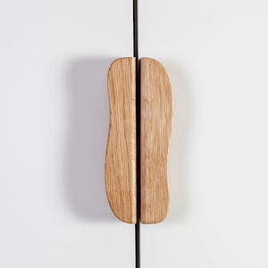 Könnte beinhalten: Zwei Holzschrankgriffe mit natürlicher Holzmaserung. Die Griffe haben eine einzigartige, organische Form und sind vertikal ausgerichtet. Sie sind an einer weißen Schranktür montiert, mit einer schwarzen Linie dazwischen.