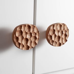 Könnte beinhalten: Zwei runde Schrankknöpfe aus Holz mit einem strukturierten, wabenartigen Muster. Die Knöpfe sind hellbraun und an einer weißen Schranktür befestigt. Die Knöpfe sind ein dekoratives Hardware-Element.