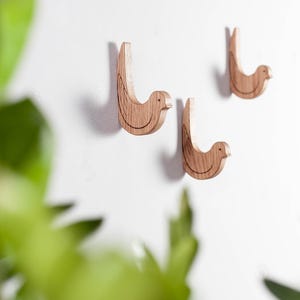 Könnte beinhalten: Drei hölzerne Vogelhaken, die an einer weißen Wand montiert sind. Die Haken sind hellbraun und haben ein schlichtes Design.