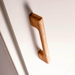 Könnte beinhalten: Ein Holz-Schrankgriff mit natürlicher Holzmaserung. Der Griff ist auf einer weißen Oberfläche montiert und wirft einen Schatten. Das Design ist modern und minimalistisch, mit einer gebogenen Form für einfaches Greifen.