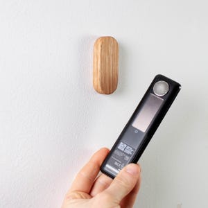May include: A black and silver electronic device with a screen and a QR code on the back. The device is being held in a hand in front of a white wall with a small wooden hook.