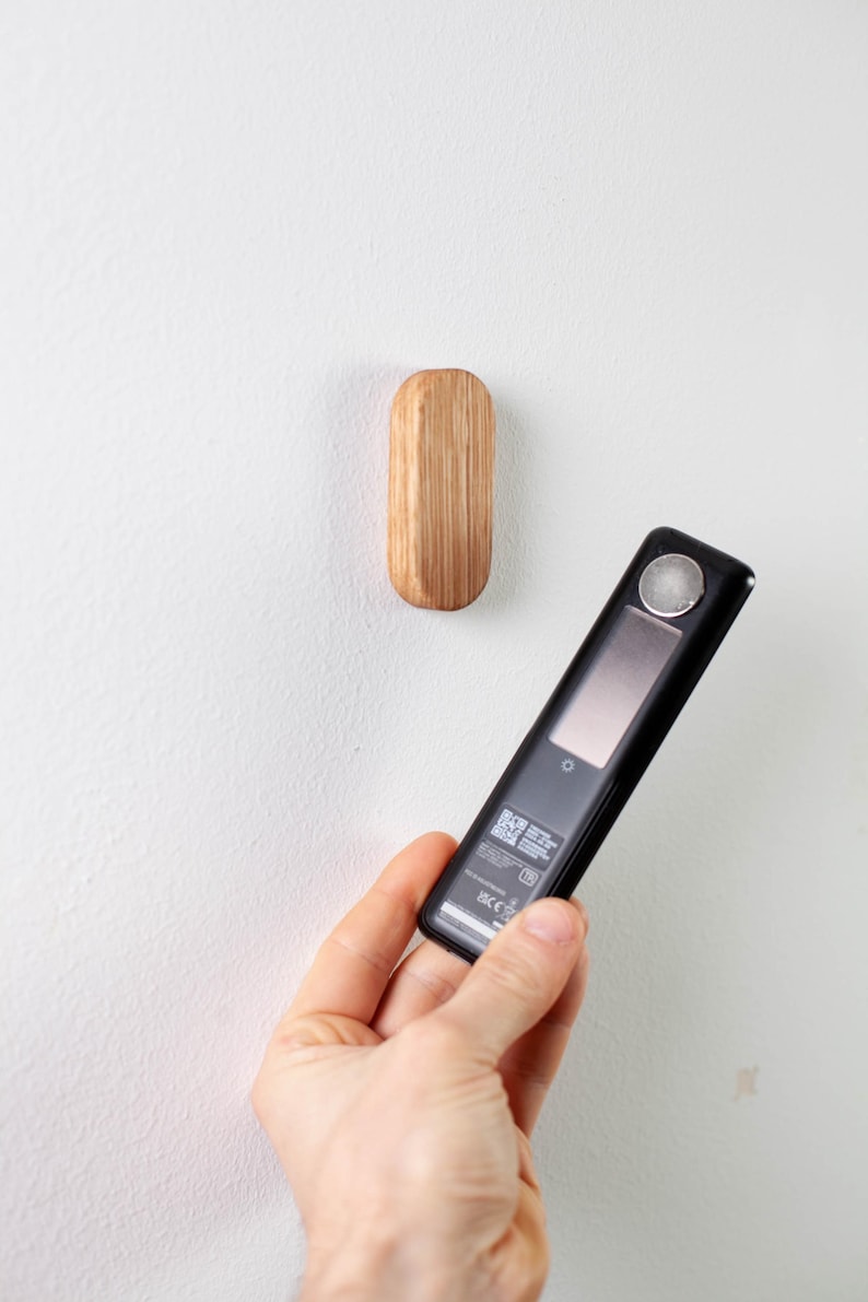 May include: A black and silver electronic device with a screen and a QR code on the back. The device is being held in a hand in front of a white wall with a small wooden hook.