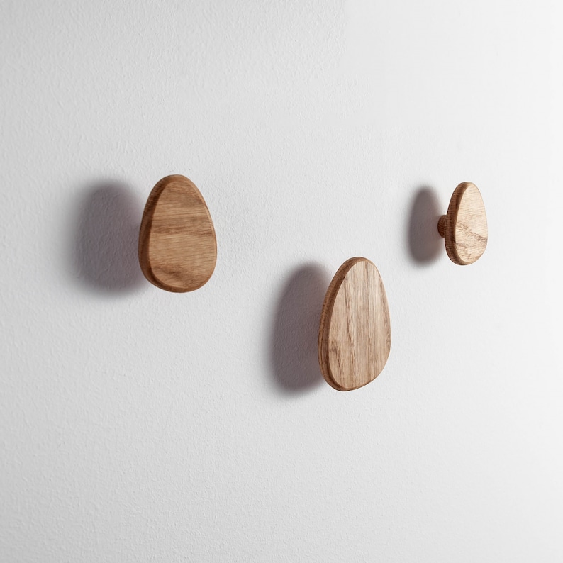 Pu&ograve; includere: Tre ganci da parete in legno a forma ovale, montati su una parete bianca. I ganci sono in legno naturale con una finitura marrone chiaro.