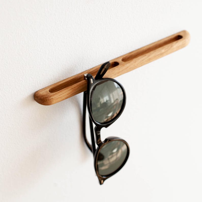 May include: A wooden wall-mounted sunglasses holder with two slots. A pair of black sunglasses is hanging from the holder.