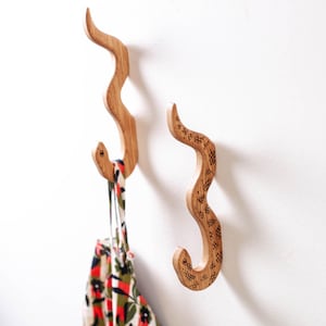 May include: Two wooden snake-shaped wall hooks. One hook has a patterned design and the other hook is holding a colourful patterned bag. The hooks are made of light-coloured wood and are mounted on a white wall.