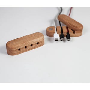 May include: Wooden cable organizers with multiple slots for cords. One organizer has cords plugged in, including a white USB-C cable, a black USB-C cable, and a black and red USB cable. The other organizer is empty.