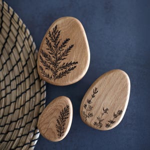Peut inclure: Trois pierres décoratives en bois de forme ovale avec des motifs botaniques gravés. Les pierres sont de couleur brun clair avec des motifs de fougères et de fleurs brun foncé. Elles sont disposées sur une surface bleu foncé.