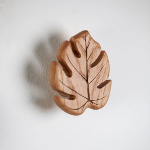 May include: A wooden leaf-shaped wall hook in a light brown colour. The leaf has a detailed carved design and is perfect for hanging items in a home or office.