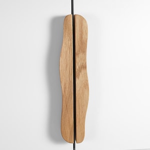 Könnte beinhalten: Zwei Holzschrankgriffe mit natürlicher Holzmaserung. Die Griffe haben eine organische Form und sind an einer weißen Schranktür montiert. Ein schwarzer Stab verbindet die beiden Griffe.