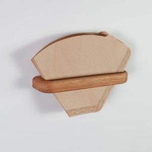 Peut inclure: Un porte-filtre à café en bois avec une pile de filtres en papier brun. Le support est en bois brun clair et possède une barre horizontale pour fixer les filtres. Les filtres sont de couleur beige clair.