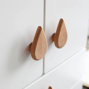 May include: Three wooden teardrop-shaped cabinet knobs in a light wood tone. The knobs are mounted on a white cabinet.