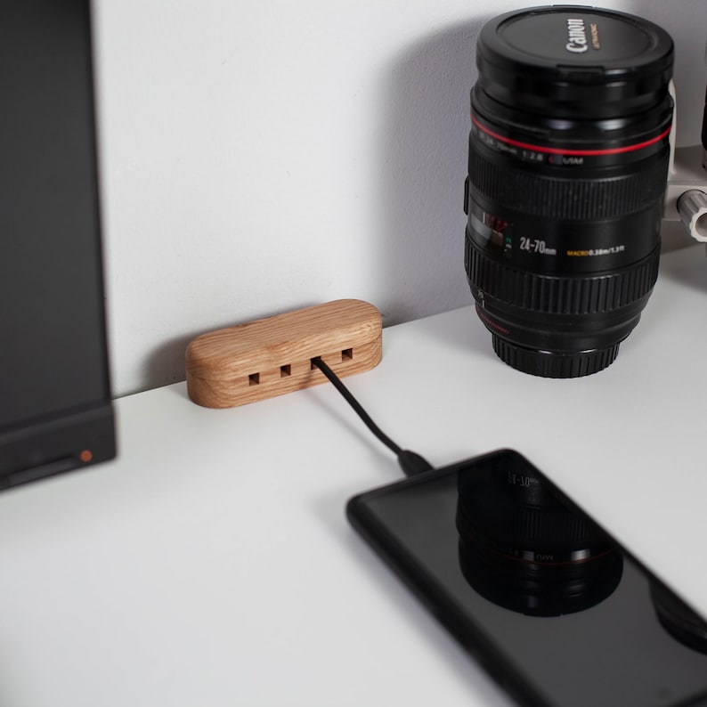 May include: A wooden charging station with three ports, connected to a black smartphone via a black cable. A black camera lens is in the background. The charging station is made of light-colored wood and sits on a white surface.