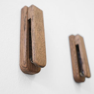 Könnte beinhalten: Zwei Wandhaken aus Holz mit natürlicher Maserung, montiert an einer weißen Wand. Die Haken haben eine rechteckige Form mit abgerundeter Oberseite und einem Metallakzent. Sie dienen zum Aufhängen von Gegenständen.