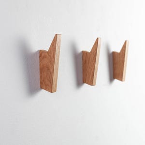 Könnte beinhalten: Drei Wandhaken aus Holz mit geometrischem Design. Die Haken sind aus hellbraunem Holz gefertigt und an einer weißen Wand montiert. Jeder Haken hat eine einzigartige, abgewinkelte Form und bietet eine moderne und minimalistische Ästhetik.
