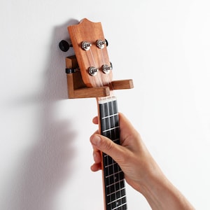 Może przedstawiać: Drewniany uchwyt ścienny na instrument, podtrzymujący główkę i gryf jasnobrązowego ukulele. Uchwyt ma wykończenie z naturalnego drewna z dwoma poziomymi ramionami wysuniętymi na zewnątrz, aby bezpiecznie podtrzymywać gryf instrumentu.