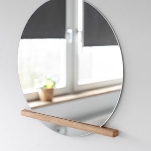 Könnte beinhalten: Ein runder Spiegel mit silbernem Rahmen und einem Holzregal, das an der Unterseite befestigt ist. Der Spiegel hängt an einer weißen Wand.