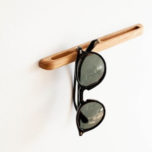 May include: A wooden wall-mounted sunglasses holder with a natural wood finish. The holder has a slot to hold the sunglasses in place. A pair of black sunglasses is hanging from the holder.