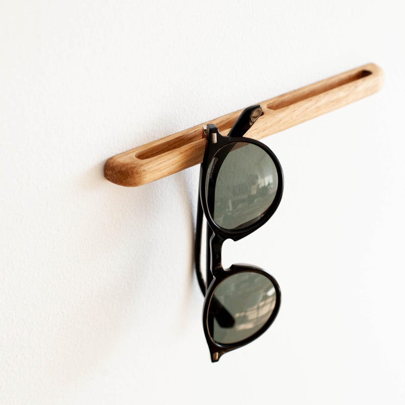 May include: A wooden wall-mounted sunglasses holder with a natural wood finish. The holder has a slot to hold the sunglasses in place. A pair of black sunglasses is hanging from the holder.