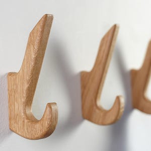 May include: Three wooden wall hooks with a natural wood grain finish are mounted on a white wall. The hooks have a curved design with a pointed top and a rounded hook. The hooks are spaced apart, creating a minimalist and functional aesthetic.