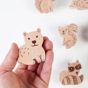 May include: Wooden animal cutouts, including a bear, squirrel, raccoon, and hedgehog. The bear is held in a hand. The animals are light brown with etched details, and are set against a white background. These wooden figures are ideal for children's toys or craft projects.