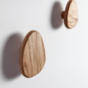 Pu&ograve; includere: Due ganci da parete in legno a forma ovale, montati su una parete bianca. I ganci sono in legno naturale di colore marrone chiaro.