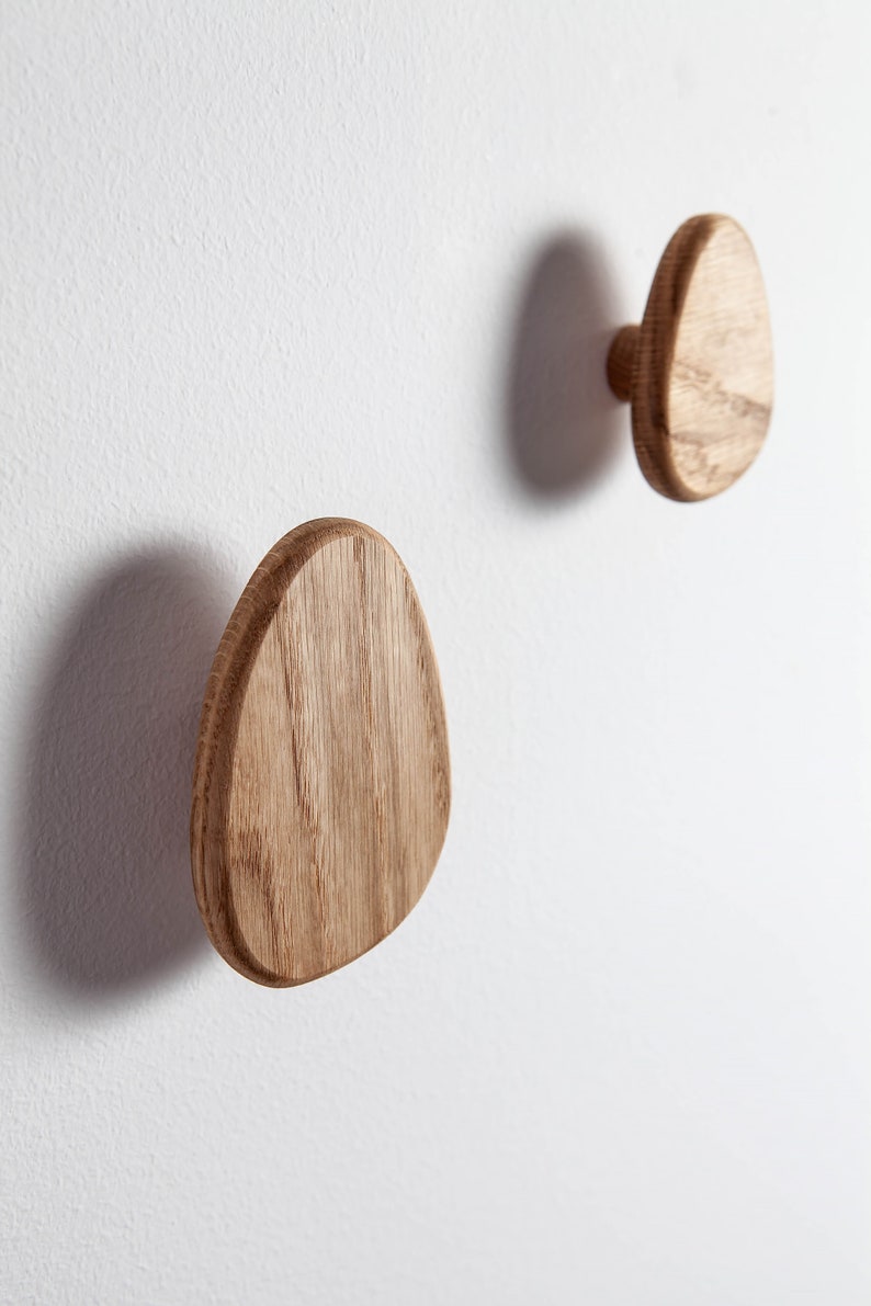 Pu&ograve; includere: Due ganci da parete in legno a forma ovale, montati su una parete bianca. I ganci sono in legno naturale di colore marrone chiaro.