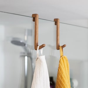 May include: Two light brown wooden hooks designed to hang over the top edge of a glass shower door. Each hook features a J-shaped profile with a flat top section and a curved bottom end. A white towel and a yellow towel are hanging from the hooks.