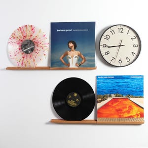Könnte beinhalten: Zwei Wandregale aus Holz mit einer transparenten Schallplatte mit rosa und gelben Sprenkeln, einem Barbara Pravi Albumcover, einer schwarzen Schallplatte, einem Red Hot Chili Peppers Californication Albumcover und einer runden Wanduhr.